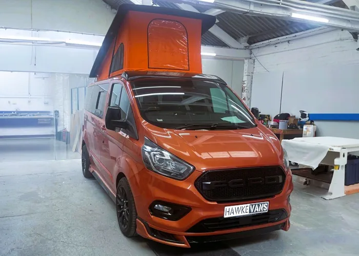 Orange camper van with a custom body kit and extended pop-top roof, parked in a workshop