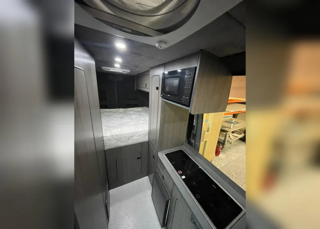 Camper van interior with a bed and compact kitchen featuring a microwave, sink, and stovetop