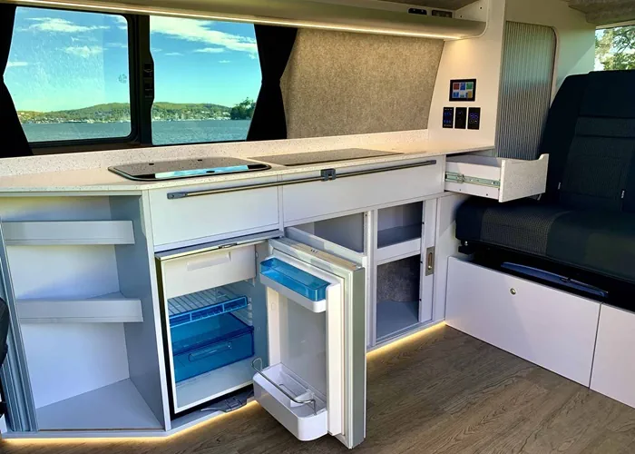 Camper van kitchenette with open storage compartments, a fridge, and a scenic view outside