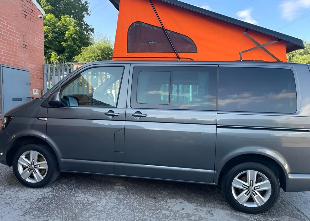 Grey camper van with an orange pop-up roof extended, parked by a brick wall