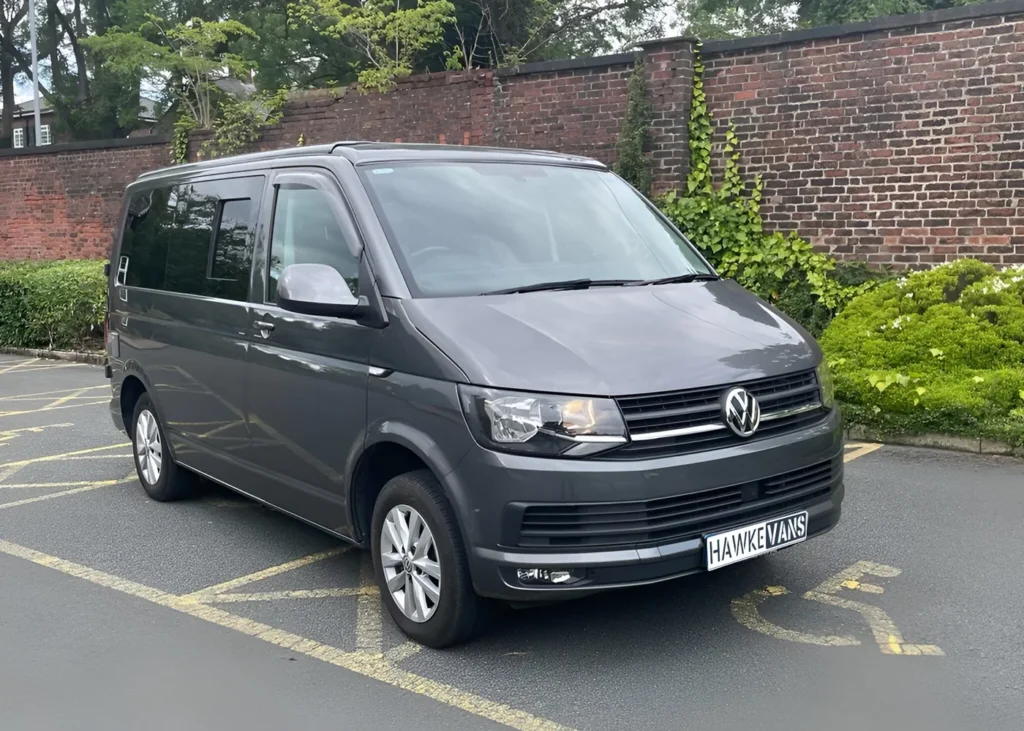 Grey camper van parked in an outdoor area with a brick wall and greenery in the background